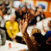 Student of color raising their hand in the foreground, in the background individuals sitting at an event listening to a speaker